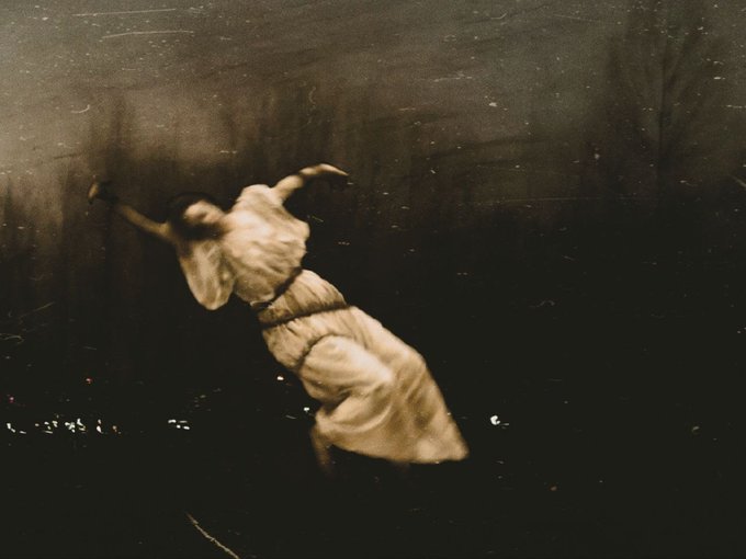 A grainy, sepia-toned photograph or film still capturing a dancer in a flowing white dress mid-leap against a dark background.