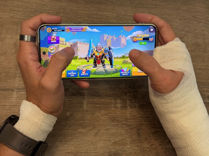 A person with bandaged hands holds a smartphone horizontally, playing a mobile game featuring a knight character with a sword on the main menu screen.