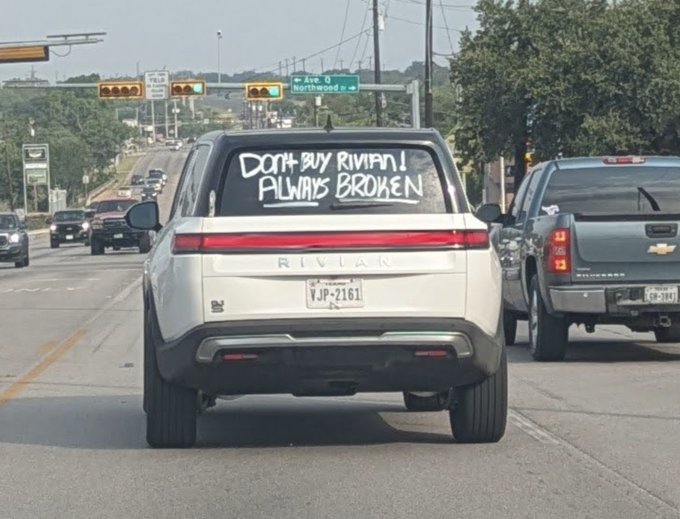 The image shows a white car with graffiti on its back window, including the words "Don't buy Riviani's broken." This message seems to be directed towards someone named Rivian, possibly an opponent or rival of the person who wrote the graffiti. The graffiti is written in black marker and appears to be spray-painted onto the car's rear window.