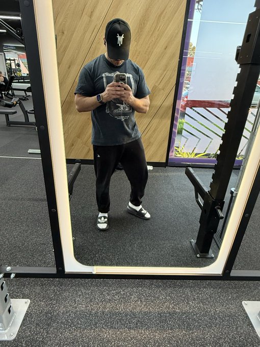 A man in a gym taking a mirror selfie while standing on a weight machine, wearing a black cap, gray t-shirt, black pants, and white/black sneakers.