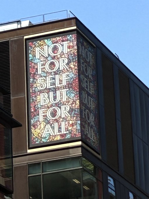 The image features a large billboard with white text that reads "not for self but for all", displayed prominently on top of a building. The billboard is situated next to a tall glass window, which allows natural light to illuminate the message. The combination of the bright white letters and the dark background creates a striking contrast, making the message stand out even more.