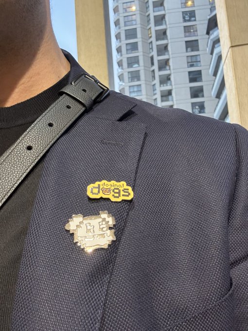 A close-up of a person wearing a dark blue blazer with two pins — a yellow "designed dogs" pin and a silver pin — with a black strap across the chest and a high-rise building in the background.