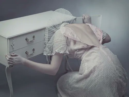An artistic photograph of a woman in a pale pink ruffled dress leaning her head against a white vintage dresser in a moody, dimly lit setting.