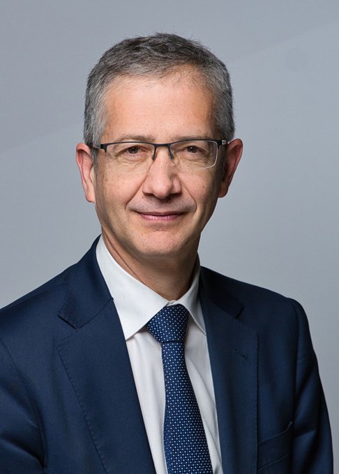 A professional portrait of a middle-aged man with gray hair and glasses, wearing a navy blue suit, white shirt, and patterned blue tie, photographed against a neutral gray background.