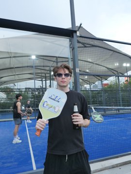 A young man wearing sunglasses on a padel court holding a paddle and a beer bottle, with other players visible in the background.
