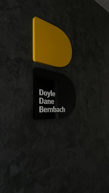 A wall-mounted sign for 'Doyle Dane Bernbach' (DDB) advertising agency featuring a yellow and black logo on a dark stone-textured wall.