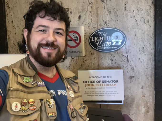 A smiling bearded man wearing a vest covered in pins stands in front of a 'Welcome to the Office of Senator John Fetterman' sign and a 'The Lighthouse Cafe 2nd Floor' sign.