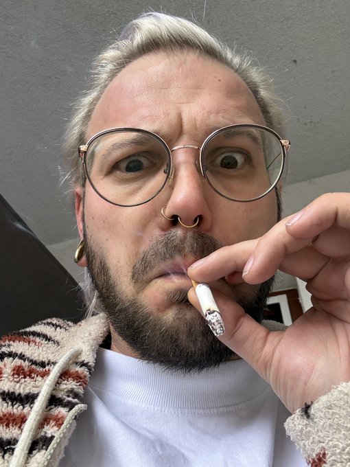 A man with blonde-streaked hair pulled back, round glasses, a beard, and a nose ring looks at the camera with wide eyes while holding a lit cigarette near his mouth.