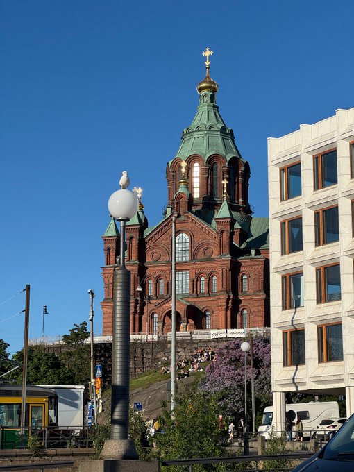 The image shows a large, old building with a green dome and a cross at its top. The building appears to be an old church or cathedral, possibly located in Helsinki, Finland. In front of the building, there are two street lamps and a traffic light. A bus is parked nearby, indicating that it's likely a busy area. There are also several people walking around, some carrying backpacks. One person has posted on social media about Bitcoin mining happening in Helsinki, suggesting an interesting connection between the old church or cathedral and the emerging world of cryptocurrency.