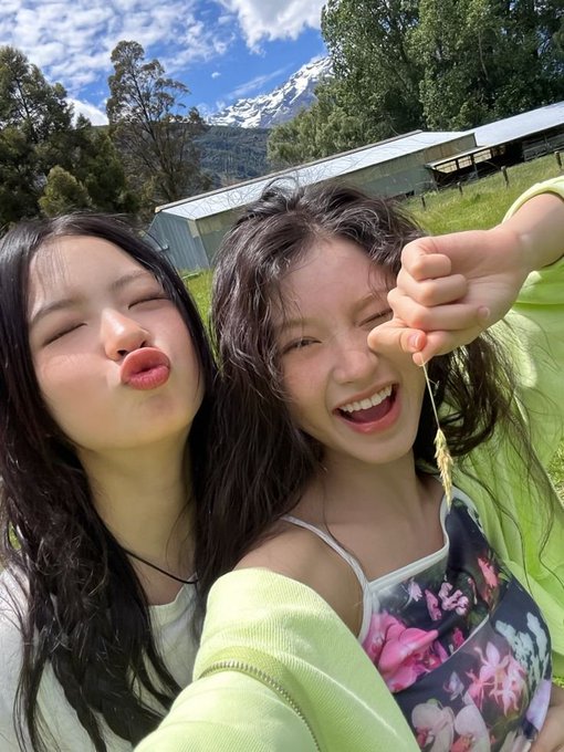 Two young women pose playfully for a selfie outdoors, with one puckering her lips and the other holding up a small plant while grinning, set against a backdrop of a snow-capped mountain, green trees, and a building under a partly cloudy blue sky.