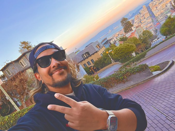 A man wearing a backwards blue cap, sunglasses, and a navy hoodie flashes a peace sign while taking a selfie, with a coastal hillside city at sunset in the background and a digital watch showing "10:05" on his wrist—capturing the friendly, sun-kissed "good morning" vibe of someone cheerfully saying "GM" to the crypto community.