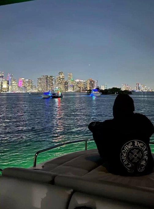 A person wearing a Chrome Hearts hoodie sits on a boat at dusk, looking out at a lit-up city skyline across the water.
