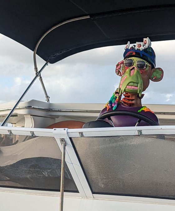 A zombie-like figure wearing sunglasses, a blue cap, and a beaded necklace sits at the helm of a boat, with colorful goop dripping down its face.