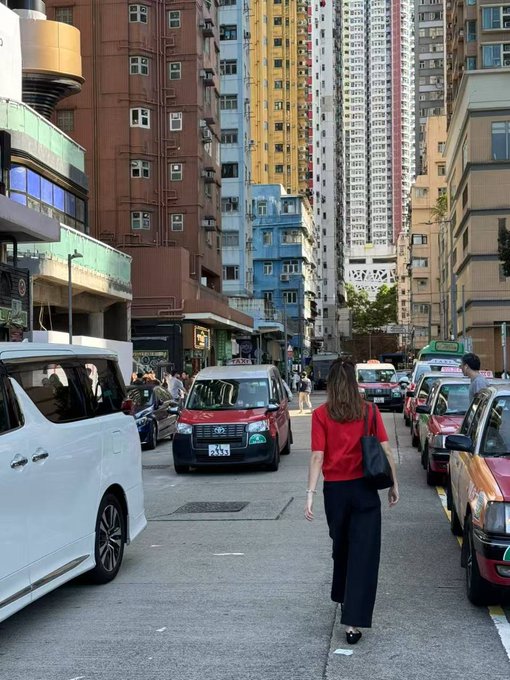 A woman in a red shirt walks down a busy city street lined with tall residential buildings and parked cars, including a van with the license plate "RJ 7335." The urban setting contrasts with the post's cautionary tale about a self-made millionaire who lost everything, suggesting themes of anonymity and the unpredictable turns of city life.