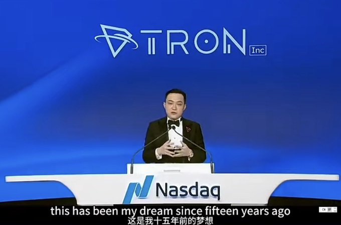 The image features a man standing at a podium with a microphone, giving a speech or presentation. He appears to be addressing an audience, possibly discussing the company's holdings structure or the platform itself. The background of the image is blue, which contrasts with the white podium and adds visual interest to the scene.