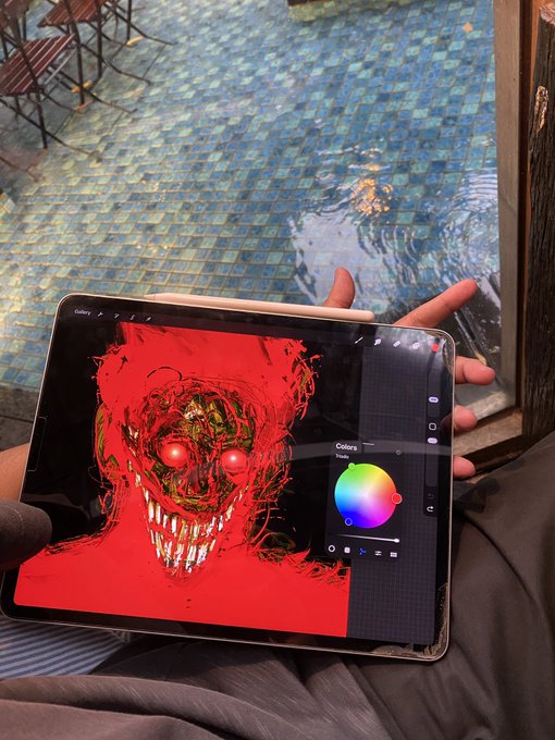A person holding an iPad displaying digital art of a sinister red-faced creature with glowing eyes in Procreate, with a color wheel visible on the right, set against a tiled patio floor.