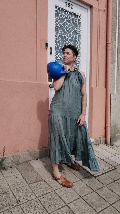 A person in a long gray tiered dress stands in front of a pink building's door (numbered 252), holding a blue balloon and a small object while wearing a dark hat.