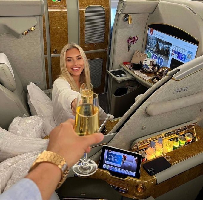 A woman in an Emirates first-class airplane suite smiling as a man's hand extends a glass of champagne toward her, with luxury amenities and a tablet visible in the cabin.