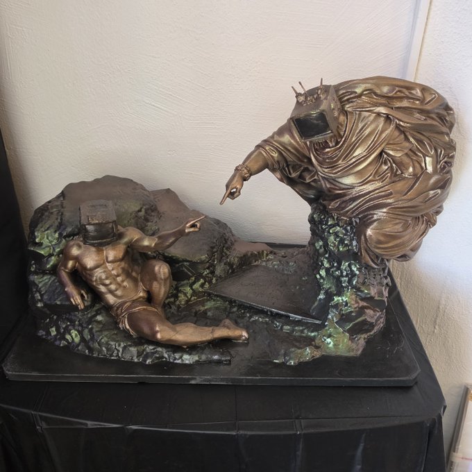 A bronze-toned fantasy sculpture shows two armored figures in a rocky scene, one reclining while pointing and the other leaning forward to point back.