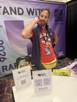 A man wearing a red Duke t-shirt and purple vest stands behind a table at what appears to be a convention booth, with QR codes and 'STAND WITH' signage visible in the background.