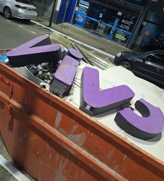 Discarded purple "Vodafone" sign letters lie broken inside a red dumpster, humorously resembling the aftermath of a late-night mining operation on the telecom company's branding.