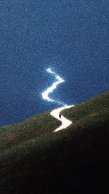 A minimalist artwork showing a small figure walking along a glowing, winding white path that ascends from a grassy hill into a dark blue sky.