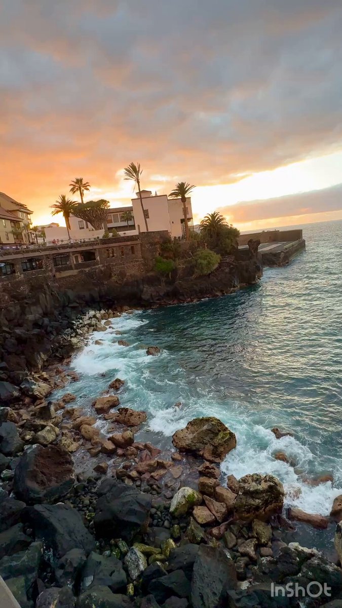 The image shows a beautiful sunset over a rocky beach, with the sky painted in shades of orange and pink as the sun sets behind the ocean. The water has small waves crashing onto the shore, creating a serene atmosphere. In the background, there is a house nestled among palm trees, adding to the tropical ambiance. A person can be seen standing on the beach, possibly enjoying the view or preparing for bedtime.
