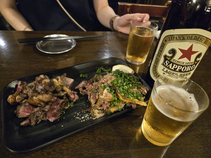 A black plate holds grilled meat topped with green onions, accompanied by a glass of beer and a bottle of 'Sapporo' lager, with a small glass of beer also visible. The label reads 'Japan's Oldest Brand Since 1876'.
