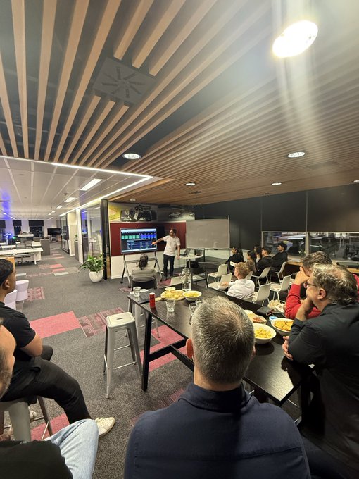 A group of people sits around a long table in a modern, well-lit room, facing a presentation screen at the front where a person appears to be speaking.