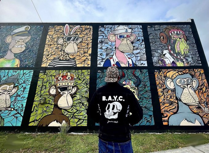 A person wearing a black jacket with 'B.A.Y.C.' and a skull design on the back stands facing a colorful mural featuring multiple cartoon monkey characters styled as Bored Ape Yacht Club NFTs.