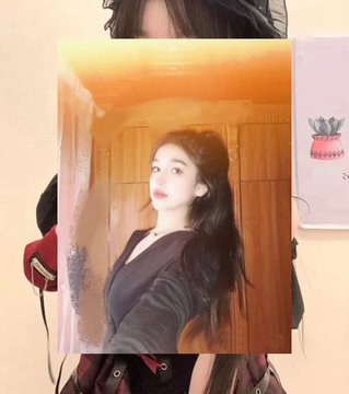 A person holds up a printed photograph of a young woman with long dark hair wearing a black top, standing in front of wooden doors with a yellow-toned background.