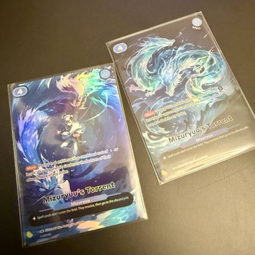 Two holographic Azuki TCG booster packs labeled "Mizuryu's Tearait" rest on a dark surface, displaying intricate dragon artwork in shimmering blues and greens, representing the alternate artwork cards mentioned in the post about a successful booster box opening.