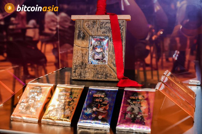 A display of ornate collectible boxes and holographic foil packs, featuring anime-style artwork and a central item adorned with a red ribbon, showcased at the Bitcoin Asia 2026 event hosted with @bis__cut. The "bitcoin asia" logo appears in the top-left corner, tying the items to the auction event.