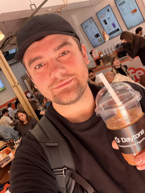 A man wearing a black cap takes a selfie while holding an iced drink from 'Daytona Computers for Agencies' in a cafe.