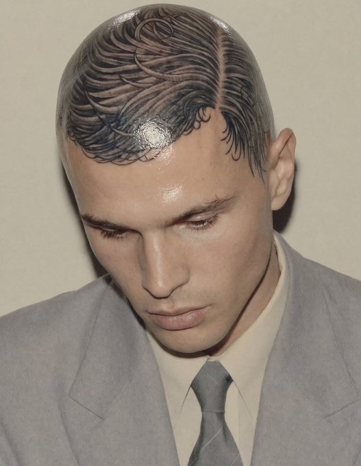 A close-up portrait of a man wearing a light grey suit and tie, looking downward, displaying a detailed haircut where the top hair is styled to resemble a feathered pattern etched into the scalp.