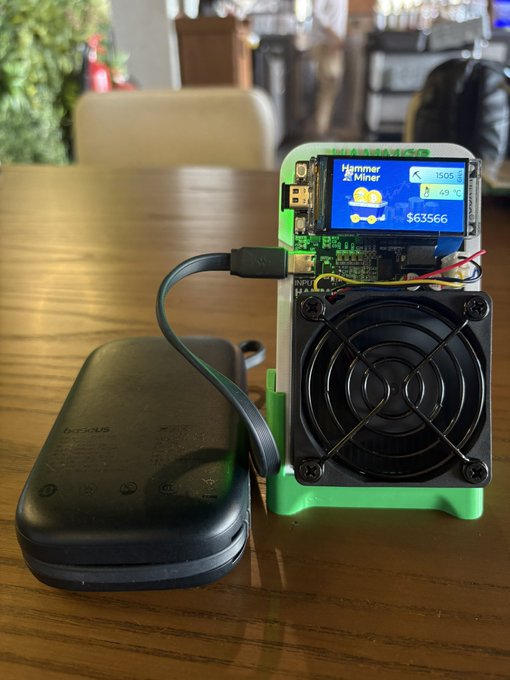A NerdMiner cryptocurrency mining device with a cooling fan and display showing 'Hammer Miner' and a balance of $63566, connected to a black power bank on a wooden table.