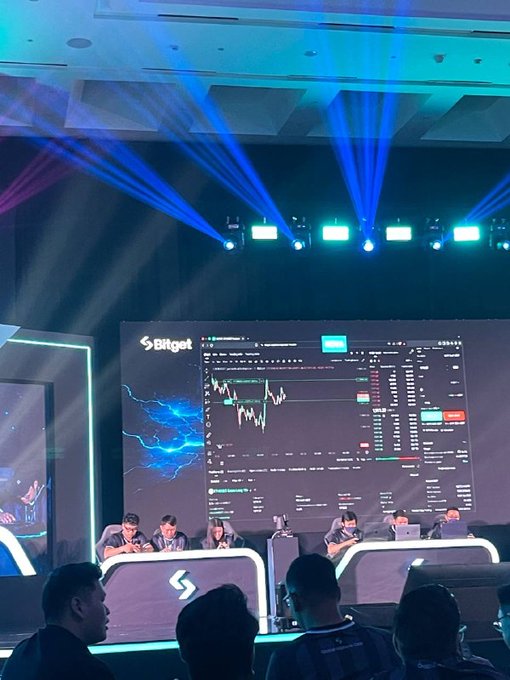 Four teams compete in the UEX Global Alpha Tournament finals on a stage featuring a Bitget-branded trading platform, with players focused on screens displaying live market charts under dramatic blue stage lighting.