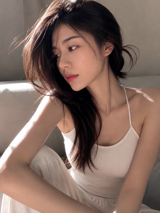 A close-up portrait of a young woman with long dark hair, wearing a white ribbed tank top, sitting on a couch in soft lighting.
