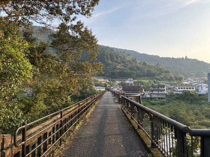 A scenic view from a bridge or elevated walkway looking toward a small town nestled among forested hills under a hazy sky.