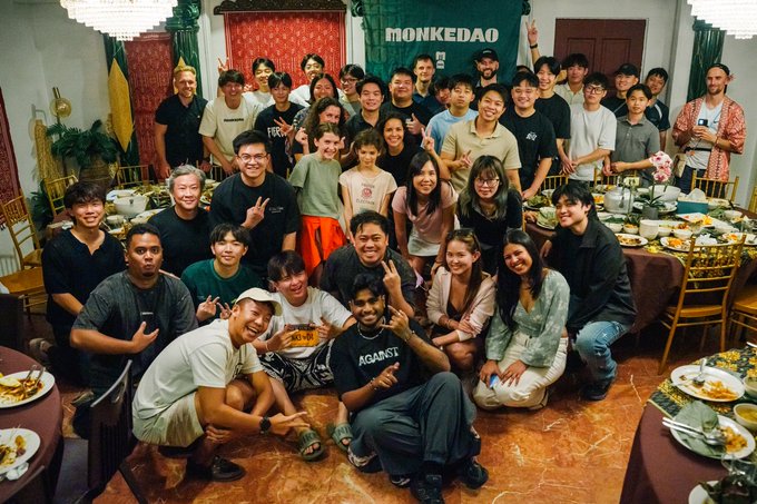 A group photo of a diverse team gathered around a dinner table, with a 'MONKEDAO' banner hanging on the wall behind them.