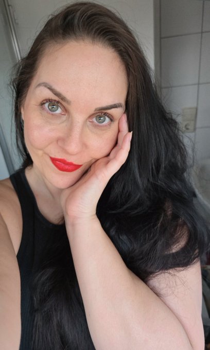 A close-up selfie shows a woman with long dark hair, striking blue eyes, and bright red lipstick resting her cheek against her hand while wearing a black tank top.
