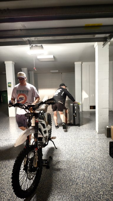 Two men are in what looks like a basement; one in a white t-shirt and cap stands near a dirt bike, while the other in black athletic wear pushes a metal cart.