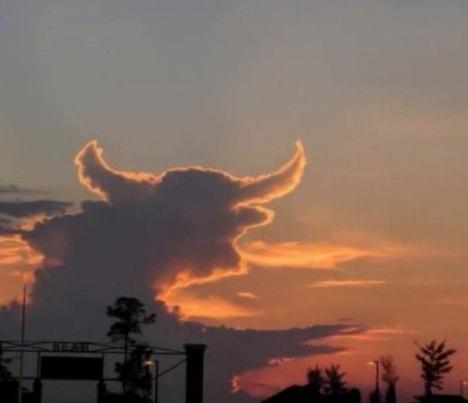 A sunset sky featuring a unique cloud formation illuminated with orange and yellow light that resembles a horned animal or devilish head shape, seen over a silhouette of trees and structures.