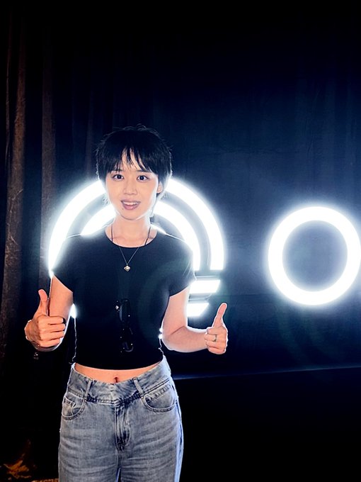 A young person with short dark hair wearing a black t-shirt and jeans gives two thumbs up while standing in front of a black background with bright circular ring lights.