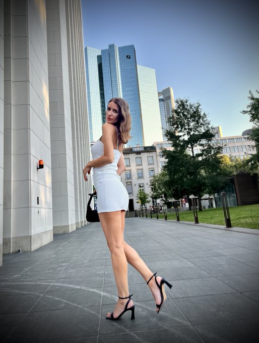 A woman in a short white dress and black high heels poses on a city sidewalk with modern skyscrapers visible in the background.