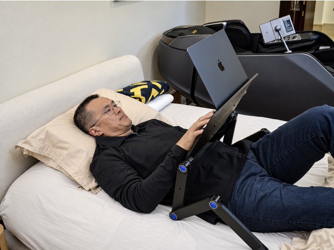A man lies back on a bed, using a laptop with a "Bitcoin" pillow nearby, humorously embodying the post's caption of making bearish $BTC tweets despite an already devastated crypto portfolio.