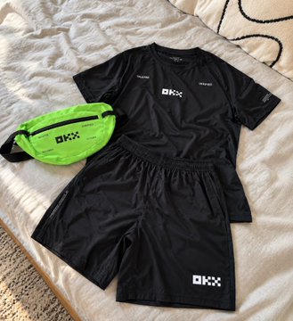 A black athletic outfit set including a t-shirt and shorts with OKX branding, paired with a neon green fanny pack, laid out on a white bed.