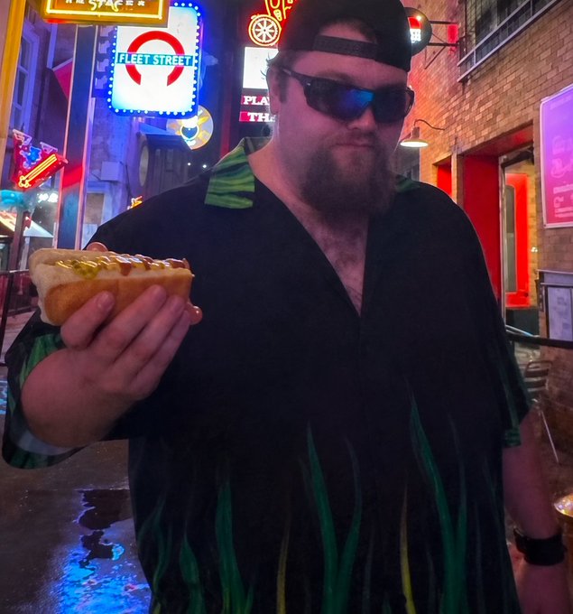 A bearded man wearing a black cap, sunglasses, and a black and green shirt holds up a hot dog on a vibrant street at night with neon signs.