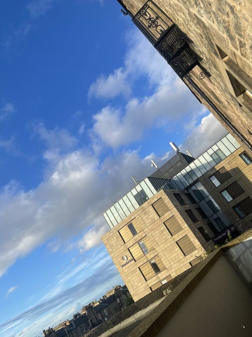 A tilted-angle photograph showing a modern building with light-colored cladding and glass windows, set against a blue sky with scattered clouds.