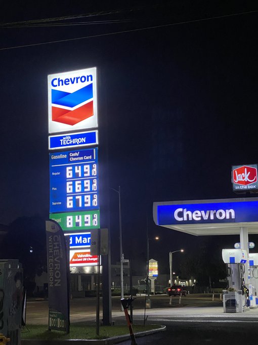 A nighttime photograph of a Chevron gas station showing fuel prices: Regular $6.49, Plus $6.69, Supreme $6.79, and Diesel $8.49, with a nearby Jack in the Box sign visible.